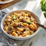 A close-up of a white bowl filled with creamy Homemade Hamburger Helper pasta, ground beef, and broccoli florets, garnished with fresh parsley on a marble surface.