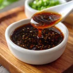 A glossy spoonful of rich brown Homemade Teriyaki Sauce with sesame seeds drips into a white bowl, with chopped green onions in the background.