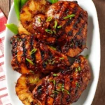 Close-up of a white plate featuring several pieces of glossy, grilled Huli Huli Chicken, garnished with green onions and served with grilled pineapple slices and green vegetables.