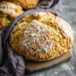 A golden-brown round loaf of Irish Soda Bread dusted with flour sits on a wooden board, with two more loaves in the background.
