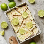 A metal pan holds various pieces of Key Lime Pie Yogurt Bark, garnished with fresh lime slices and zest, surrounded by whole and halved limes on a textured gray background.