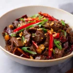 A close-up shot of a bowl filled with delicious Kung Pao Beef, garnished with bright red chilies, green onions, and chopped nuts.