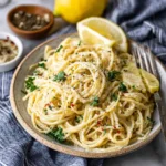 A vibrant plate of creamy Lemon Spaghetti garnished with fresh parsley, black pepper, and red pepper flakes, with lemons in the background.