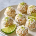 A plate filled with round Lime Pie Energy Bites, coated in shredded coconut and garnished with lime zest, alongside fresh lime wedges.