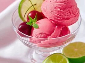 Vibrant pink Ninja Creami Cherry Limeade Sorbet scoops in a glass bowl, garnished with cherries, lime, and mint, with lime halves nearby.