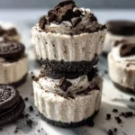 Close-up of two stacked no-bake Oreo cheesecake bites, garnished with whipped cream and crushed Oreos, on a marble counter.