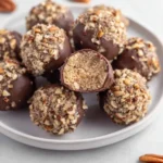 A white plate piled high with chocolate-dipped No Bake Pecan Pie Balls, most coated in chopped pecans, with one cut open revealing the crumbly filling.