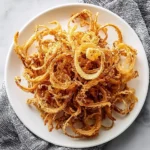 A white plate filled with a large serving of golden crispy fried Onion Strings, set on a white marble surface with a gray patterned napkin.
