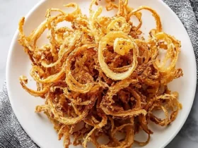 A white plate filled with a large serving of golden crispy fried Onion Strings, set on a white marble surface with a gray patterned napkin.