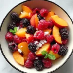 A vibrant Peach Berry Fruit Salad in a white bowl, featuring fresh peaches, raspberries, and blackberries, garnished with mint.