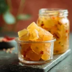 A small glass bowl and a jar brimming with glistening diced Pineapple Cowboy Candy, featuring visible chili flakes, on a dark countertop.