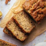 A freshly baked loaf of Pineapple Zucchini Bread, partially sliced, rests on parchment paper next to cinnamon sticks and star anise.