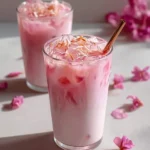 Two glasses of vibrant Pink Coconut Iced Latte with ice and delicate flower petals, surrounded by scattered pink blossoms.