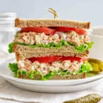 A tall, delicious salmon salad sandwich on whole wheat bread, filled with creamy salmon, lettuce, and tomato, served with pickles.