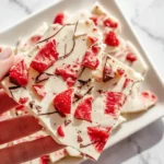 A hand holds a piece of creamy white strawberry yogurt bark, adorned with vibrant red dried strawberries and dark chocolate flecks.