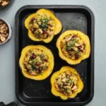 Four golden Stuffed Acorn Squash halves filled with quinoa, mushrooms, cashews, and pomegranate seeds on a black baking tray.