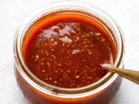 A glass jar filled with rich, homemade taco sauce, featuring a spoon dipped into it on a light textured background.