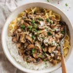 A top-down view of a creamy Vegan Mushroom Stroganoff served over white rice and egg noodles, garnished with fresh parsley in a white bowl.