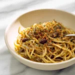 A close-up of a bowl of delicious Anchovy Pasta with Breadcrumbs, garnished with fresh herbs, sitting on a marble table.
