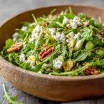 A vibrant Arugula Goat Cheese Salad in a rustic wooden bowl, garnished with pecans, grapes, and seeds.
