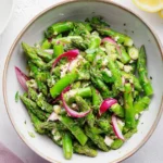 A vibrant bowl of fresh asparagus salad tossed with thinly sliced red onions, crumbled feta cheese, and fresh herbs, with a lemon half visible.