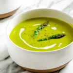 A close-up of a white ceramic bowl filled with vibrant green asparagus soup, garnished with two asparagus tips, a cream swirl, and black pepper on a marble surface.