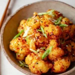 A close-up of crispy, golden bang bang cauliflower florets garnished with black and white sesame seeds and green onions in a textured bowl.