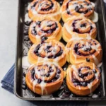 Eight warm blueberry cinnamon rolls, generously drizzled with white icing, in a black baking pan.