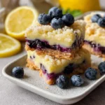 Two stacked Blueberry Lemon Bars with crumbly topping, creamy lemon layer, and blueberry filling, garnished with fresh blueberries and lemons in the background.