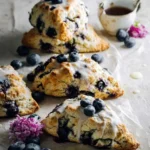 Several homemade Blueberry Sourdough Scones with glaze and fresh blueberries, served on parchment paper with a small teacup.