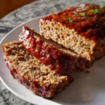 A freshly baked Brown Sugar Meatloaf, beautifully glazed and garnished with herbs, with two slices cut and served on a white plate.
