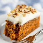 A delicious slice of Carrot Cake with Cream Cheese Frosting, topped with chopped nuts, on a white plate with a fork.