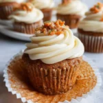 A delicious carrot cupcake with creamy white frosting and a topping of chopped carrots and nuts, with more carrot cupcakes blurred in the background.