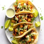 Four delicious Cauliflower Tacos with Cilantro Lime Crema on a white plate, garnished with fresh cilantro and lime wedges.