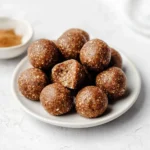 A white plate holds numerous brown Chai Spice Protein Balls, one broken to show its textured interior, with a spice bowl nearby.