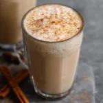 A steaming Chai Tea Latte in a clear glass mug, topped with frothy foam and a sprinkle of cinnamon, with cinnamon sticks nearby.