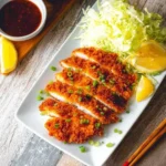 Sliced crispy golden Chicken Katsu with shredded cabbage, lemon wedges, a side of rice, and tonkatsu sauce.