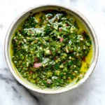 A vibrant green Chimichurri Sauce with chopped herbs, red onion, and olive oil in a rustic bowl on marble.