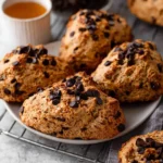 A plate of freshly baked Chocolate Chip Coffee Scones, rich with chocolate chips, sits on a cooling rack beside a mug of coffee.