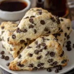 A close-up shot of several homemade chocolate chip scones piled on a white plate with coffee in the background.