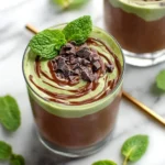 A refreshing Chocolate Mint Smoothie in a glass, layered with chocolate and mint cream, adorned with chocolate chunks and fresh mint.