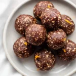 A close-up view of a pile of textured chocolate orange bliss balls on a light grey plate, garnished with flecks of orange zest.