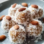 A close-up of several homemade Coconut Date balls coated in shredded coconut and topped with an almond, arranged on a grey plate.