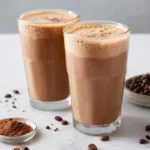 Two glasses of frothy Coffee Protein Smoothie with chocolate shavings, coffee beans, and cocoa powder on a marble surface.