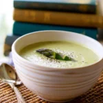 A creamy bowl of Cream of Asparagus Soup garnished with fresh asparagus spears and black pepper, next to an antique spoon and stacked books.