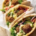 Three vibrant Falafel Pita Sandwiches, filled with crispy falafel, fresh lettuce, cucumber, tomato, red onion, and creamy sauce, served in white paper.