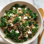 A close-up view of a vibrant Farro Kale Salad With Goat Cheese, also featuring raisins and pumpkin seeds, in a white bowl.