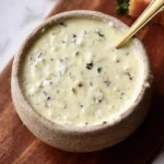 A rustic bowl filled with creamy Garlic Parmesan Sauce, garnished with green herbs, with a golden spoon dipping in. Served on a wooden board with bread and garlic.