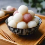 A striped bowl brimming with white and pink glutinous rice balls, presented on a wooden tray alongside chopsticks.