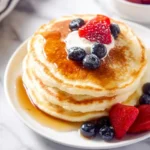 A tempting stack of fluffy greek yogurt pancakes drizzled with maple syrup, topped with fresh strawberries, blueberries, and whipped cream.